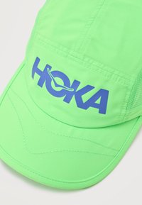 Φωτεινό πράσινο ελαφρύ καπέλο με μπλε λογότυπο HOKA και διχτυωτά πλαϊνά πάνελ, σχεδιασμένο για αθλητική χρήση.