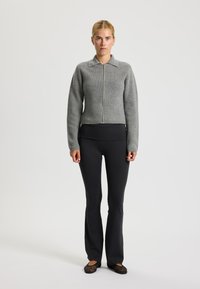 Grijze geribbelde zip-up trui met een kraag, gecombineerd met zwarte aansluitende broek en zwarte platte schoenen. De outfit is eenvoudig en aansluitend.