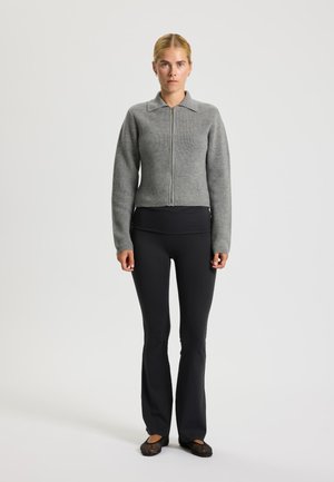 Maglione grigio a coste con zip e colletto, abbinato a pantaloni neri aderenti e scarpe flat nere. L'outfit è semplice e aderente.