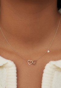 Halskette in Goldton mit zwei miteinander verbundenen Herzformen und einem kleinen Herzanhänger, vor einem strukturierten Hauthintergrund.