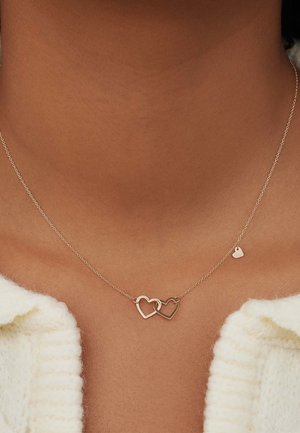 Gold-tone necklace featuring two interlocking heart shapes and a small heart pendant, set against a textured skin background.