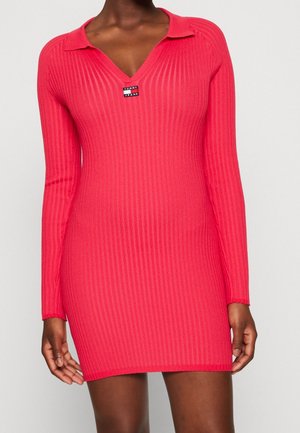 Frau trägt ein langärmliges, geripptes, hellpinkes Strickkleid mit Kragen und einem kleinen "Tommy Jeans"-Logo auf der Brust.