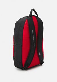 Svart och röd ryggsäck med vadderad design, justerbara svarta axelremmar, sidoficka och en synlig slogan tryckt på remmen.