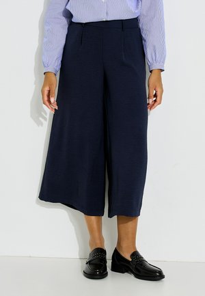 Persona que lleva pantalones anchos azul marino recortados, blusa a rayas azul y blanca, y mocasines de cuero negro con pequeñas tachuelas.