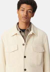 Crème korduroy jack met knoopsluiting, twee voorkockets, een puntige kraag en een gestructureerd oppervlak. Draag over een geruite blouse.