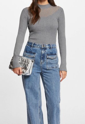 Femme portant un pull gris moulant en maille côtelée avec un col rayé transparent et un jean bleu taille haute, tenant une pochette matelassée argentée.