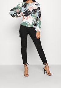 Blusa multicolore con maniche a sbuffo lunghe e motivi floreali; abbinata a pantaloni neri slim-fit e tacchi alti neri con cinturini.