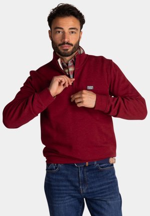 Rode fleece pullover met een ritssluiting bij de kraag, voorzien van een klein logopatch op de borst. Het is gedragen over een geruite shirt en gecombineerd met blauwe jeans.