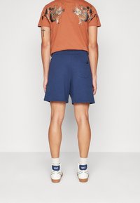 Shorts bleu marine avec une taille élastique, des poches latérales et une poche arrière. Porté avec un T-shirt marron arborant des détails graphiques.
