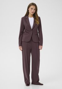 Bordeauxroter maßgeschneiderter Anzug mit einem taillierten Blazer, der über zwei vordere Taschen verfügt, und weitem Hosenbeinen. Getragen über einem weißen Hemd. Glatte Stofftextur.
