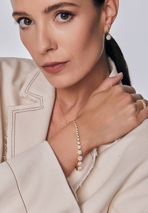 Złota bransoletka z naprzemiennymi przezroczystymi kamieniami, noszona na nagiej skórze. W tle widoczna kremowa marynarka z postrzępionymi brzegami.