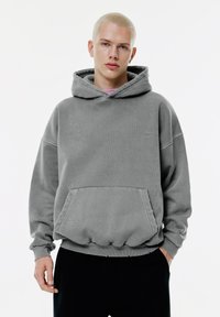 Felpa grigia con cappuccio, tasca frontale, polsini e orlo a coste. Tessuto morbido con un leggero motivo tonale. Indossata con pantaloni neri.