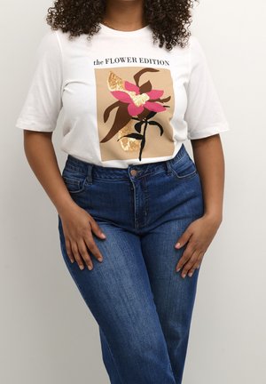 Vrouw die een wit T-shirt draagt met een roze en gouden bloemenontwerp met pailletten en de tekst "the FLOWER EDITION", gecombineerd met een blauwe spijkerbroek.