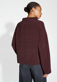 Bordeaux oversized pullover met een textuurafwerking, voorzien van brede mouwen en een hoge hals, gedragen met donkere denimbroeken.