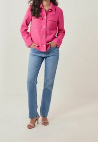 Veste rose texturée avec deux poches poitrine et boutons argentés, associée à un jean droit bleu clair et des sandales à talons nus.