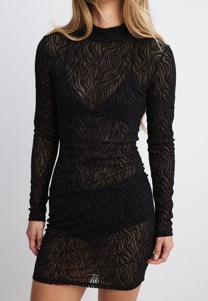 Femme portant une robe noire moulante à manches longues en tissu transparent avec un motif subtil de rayures ondulées, debout devant un fond uni.