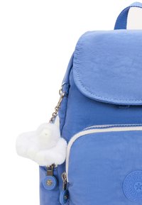 Modrý nylonový batoh s kulatým klopou, bílými detaily, kapsami na zip a bílým chlupatým klíčenkou ve tvaru opice připevněnou na stříbrném klipsu.