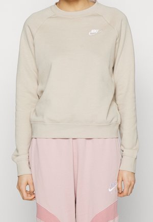 Person iført beige Nike sweatshirt og lyserøde Nike Air joggingbukser, stående mod en ensfarvet baggrund.