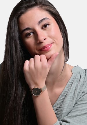 Young woman with long dark hair wearing a wristwatch, resting her chin on her hand, and smiling softly against a plain background.