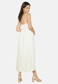 Vestido maxi blanco con una delicada textura bordada, tirantes finos y cintura ceñida. Sandalias doradas de tacón cuadrado complementan el conjunto.
