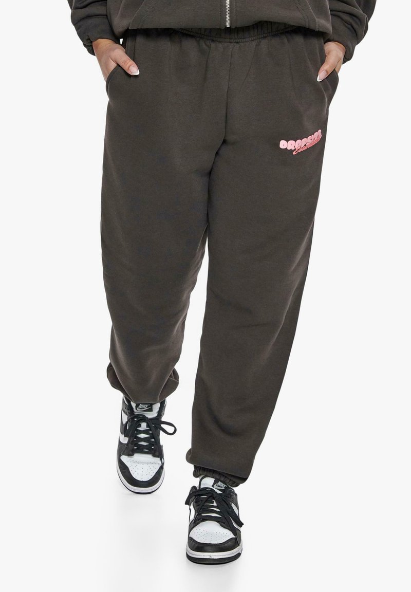 Dunkelgraue Sweatpants mit elastischem Bund, Seitentaschen und einem rosa grafischen Design auf dem Oberschenkel. Getragen mit schwarz-weißen Sneakern.
