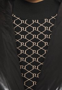 Černá péřová bunda s lesklou texturou, pod kterou se nachází černý a růžový svetr s geometrickým vzorem.