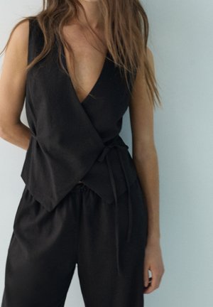 Femme portant un haut cache-cœur noir sans manches et un pantalon noir ample assorti sur un fond uni clair.