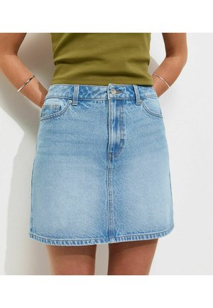 Vrouw draagt een lichtblauwe spijkerrok en een olijfgroene mouwloze top, handen rustend achter haar rug, tegen een witte achtergrond.