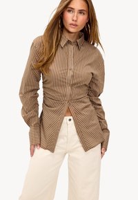 Brown and white striped button-up shirt with a fitted design and gathered waist, paired with light beige wide-leg pants.