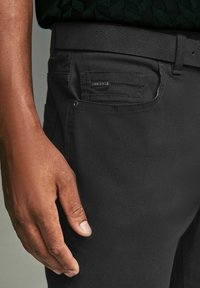 Pantaloni negri dintr-un material texturat, cu un buzunar lateral ce poartă o etichetă. O mână se sprijină pe coapsă, evidențiind ajustajul și detaliile de design.