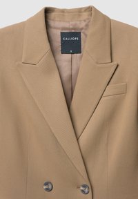 Blazer beige à double boutonnage avec revers, deux boutons et une poche poitrine. Tissu lisse et texturé ; l'étiquette au col indique "CALLIOPE."