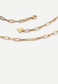 Gouden ketting met verlengde schakels, een driehoekige hanger met het "GUESS" logo en een karabijnhaaksluiting.