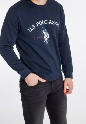 Mężczyzna ubrany w granatową bluzę U.S. Polo Assn. z białym logo i napisem, w połączeniu z czarnymi jeansami, ręka w kieszeni, na białym tle.