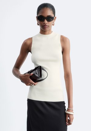Femme portant des lunettes de soleil noires, un haut blanc sans manches, une jupe noire, tenant un sac à main noir, avec des boucles d'oreilles et des bracelets sur un fond clair.