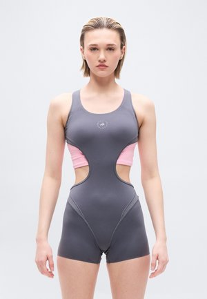 Joven con cabello corto y rubio que lleva un body deportivo gris y rosa con recortes, de pie contra un fondo claro y liso.