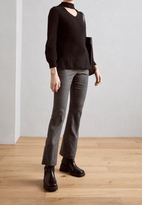 Osoba ubrana w czarny golf z chokerem, szare spodnie w wzory, czarne botki oraz niosąca czarną torbę na ramię na drewnianej podłodze.