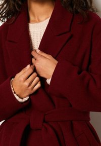 Femme portant un manteau ceinturé rouge profond par-dessus un pull en tricot rose clair, ajustant le manteau près du col avec les deux mains.