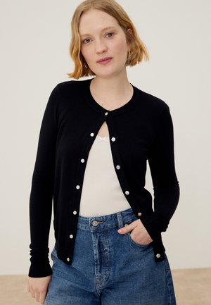 Νέα γυναίκα με κοντά ξανθά μαλλιά φοράει μια μαύρη ζακέτα με κουμπιά πάνω από ένα λευκό τοπ και μπλε τζιν, στέκεται με το ένα χέρι στην τσέπη.