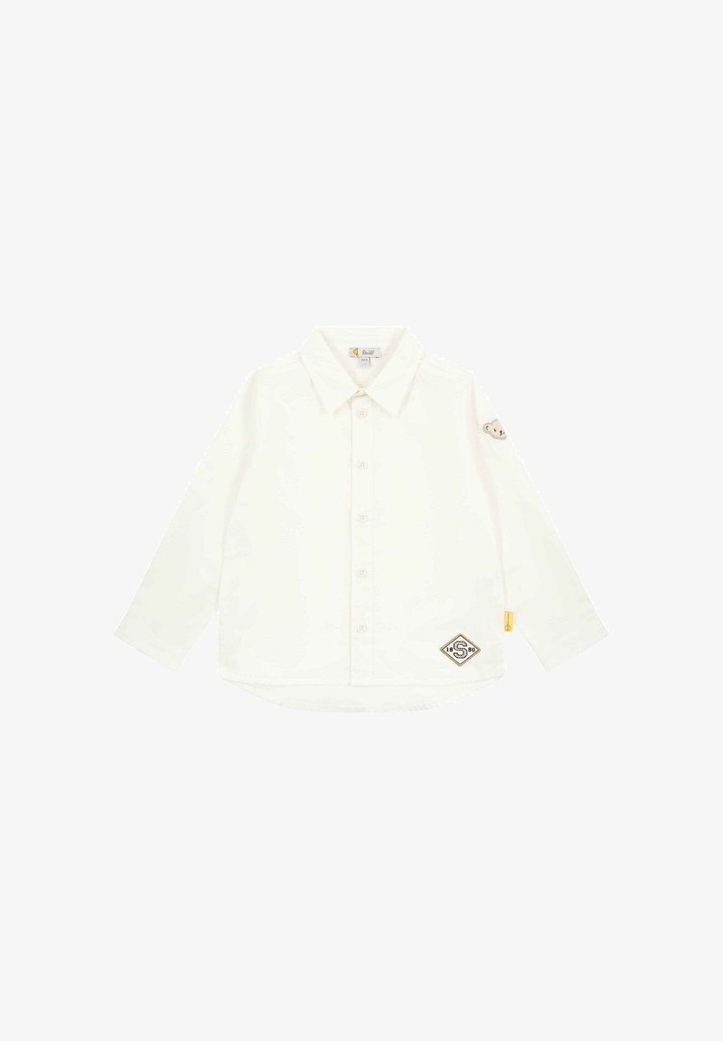 Biała koszula z długim rękawem, zapinana na guzik, z kołnierzykiem, z brązową łatą od misia na rękawie oraz logo na dole. Miękka tekstura.