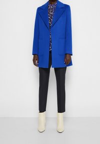 Abrigo de mezcla de lana azul con un amplio cuello y bolsillos frontales, combinado con una camisa floral, pantalones negros ajustados y botas de tobillo crema.