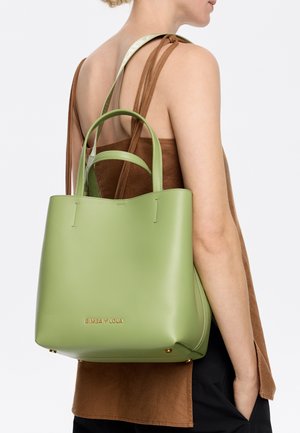 Green leather tote bag with a minimalist design, featuring dual handles and gold logo detail on the front, positioned on a model's shoulder.