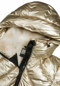 Guldmetalisk pufferjakke med en blød hvid pelsfoering, med en sort lynlås og hætte samt et logomærke indeni.