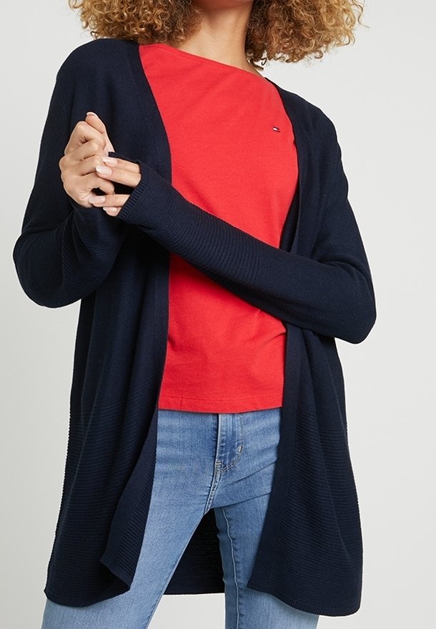Femme portant un cardigan bleu marine sur une chemise rouge et un jean bleu clair, les bras croisés à la taille.