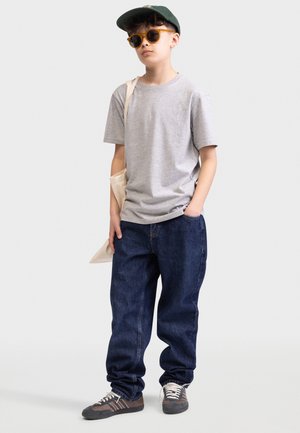 T-shirt en coton gris, jean en denim bleu foncé ample, chaussures grises et brunes, lunettes de soleil jaunes, casquette verte, sac fourre-tout crème.