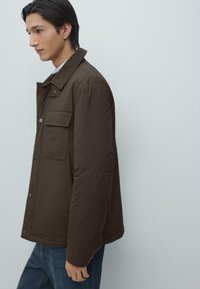 Brun vadderad jacka med krage, utrustad med en bröstficka och tryckknappar. Bärs med blå jeans, visas från sidan.