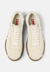 Sneaker in tela beige con bordi sfrangiati, lacci bianchi e suola in gomma marrone. L'interno presenta un testo di logo rosso su sfondo crema.