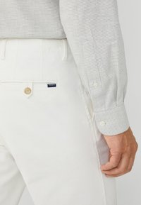 Pantaloni bianchi in cotone con una tasca posteriore con bottone e un'etichetta blu navy. Una camicia a scacchi grigio chiaro presenta polsini con bottone.