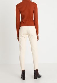 Pull à col roulé côtelé de couleur rouille associé à un jean ajusté ivoire avec des ourlets effilochés, complété par des bottines noires.