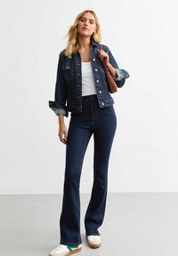 Jeans acampanados de mezclilla oscura combinados con una chaqueta de mezclilla azul clara. La modelo lleva una blusa blanca y sostiene un bolso marrón. Las deportivas tienen acentos verdes.