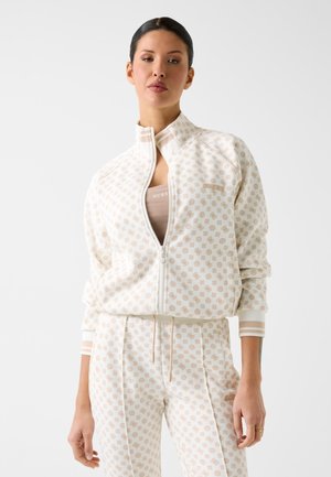 Femme portant une veste blanche zippée et un pantalon assorti avec un motif beige répété, debout devant un fond clair uni.